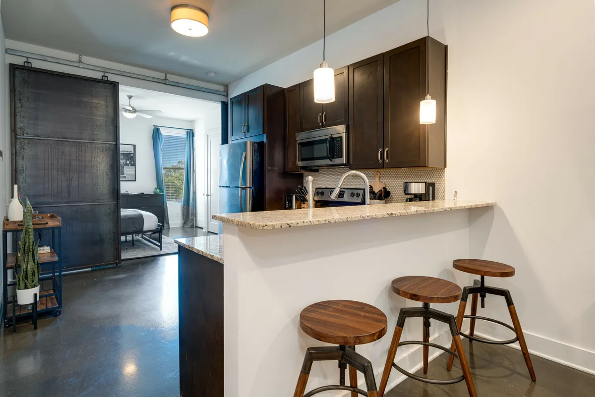 A modern kitchen scene featuring a sleek granite countertop with a set of bar stools, contemporary kitchen appliances, and a sliding barn door in the background. The space feels open and perfect for both cooking and socializing, with clean lines and a bright, inviting atmosphere.