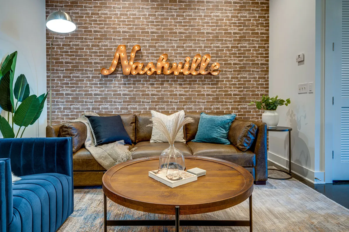 A chic living area featuring a brown leather sofa, a blue accent chair, a Nashville-themed sign, and a stylish coffee table. Contemporary artwork and modern decor set a warm, inviting tone, perfect for social gatherings or relaxing after a day of exploring.