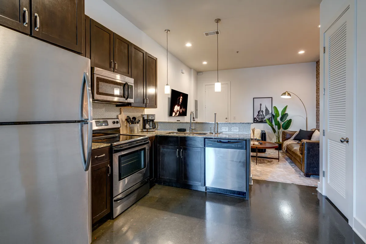 A bright, contemporary loft interior featuring stainless steel appliances, a kitchen island, a comfortable living room seating area, and modern art on the wall. The scene is welcoming and organized, reflecting a well-maintained and professionally managed property.