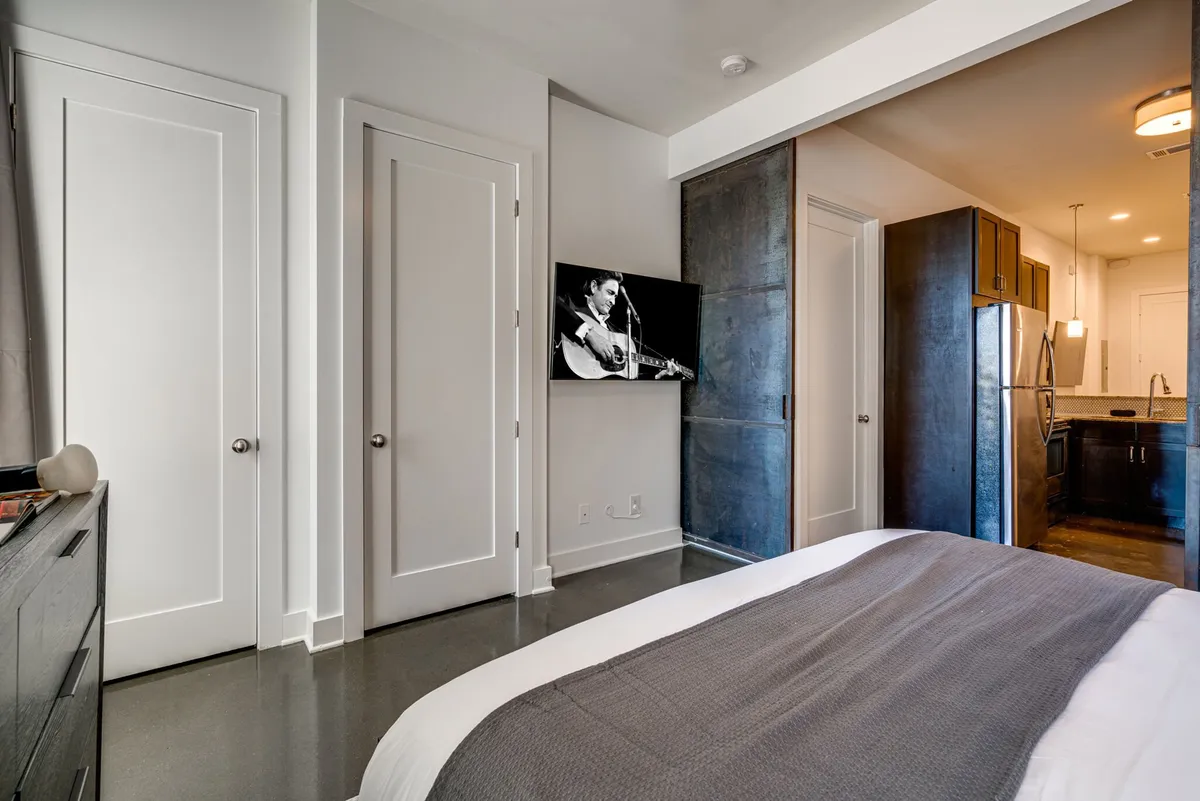A modern bedroom featuring a comfortable bed with gray bedding, a wall-mounted flat-screen TV showing a musician, and sliding closet doors. The atmosphere is contemporary and inviting, suggesting options for entertainment and relaxation.