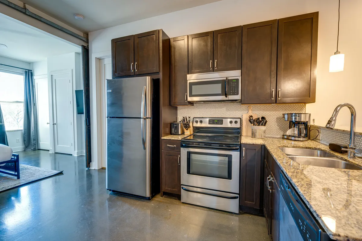 A cozy kitchen area featuring stainless steel appliances, ample granite countertops, and dark wooden cabinets. Natural light pours in through a window, illuminating the space and making it ideal for preparing meals or enjoying a quiet coffee moment.
