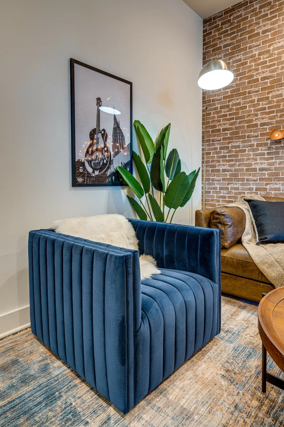 A stylish living space featuring a blue velvet chair, a brown leather sofa, framed artwork, a green plant, and a wooden coffee table. The decor blends comfort with artistic inspiration, ideal for both socializing and quiet moments.