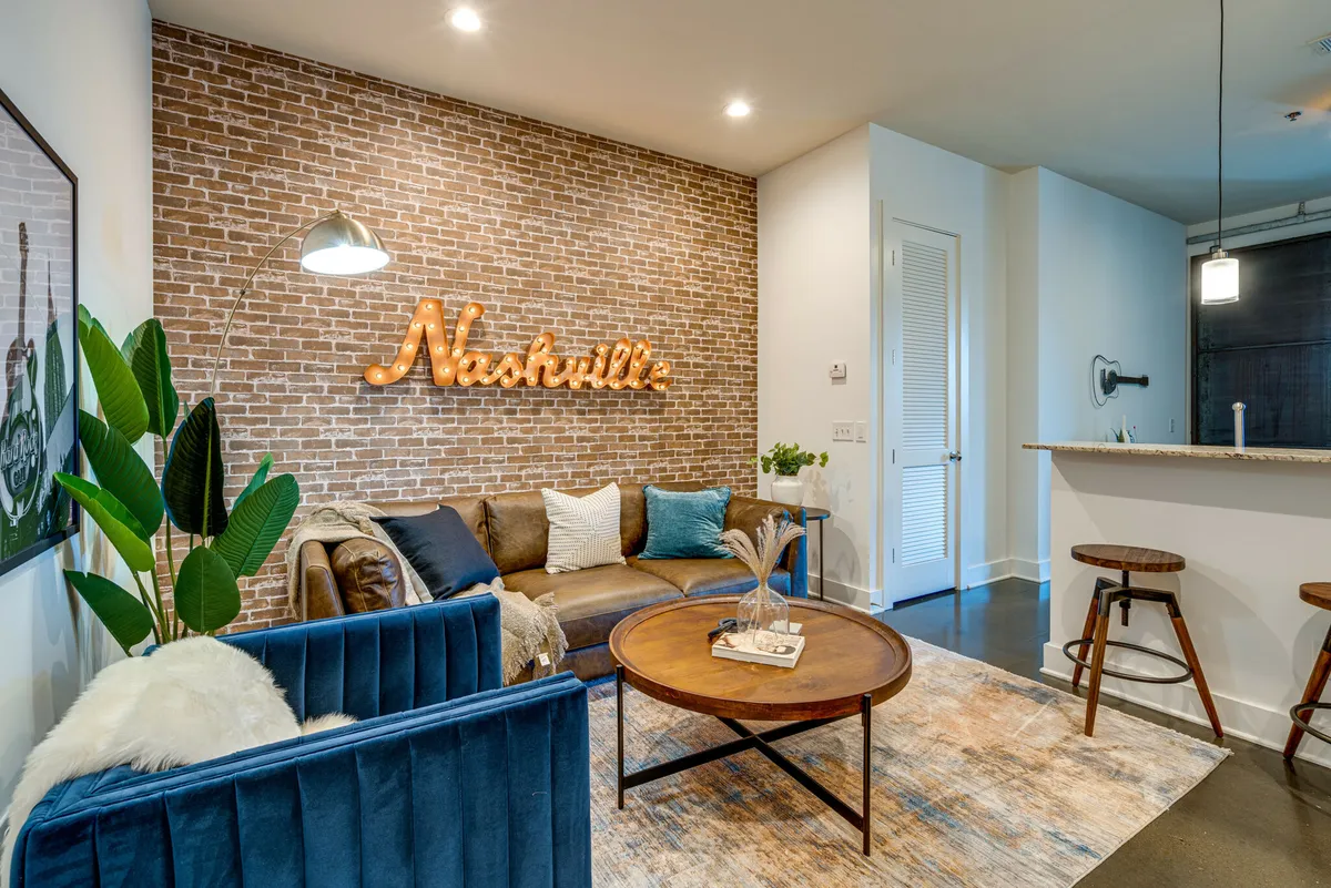 The image shows a stylish, cozy living area with exposed brick walls featuring a bold 'Nashville's' sign, a rich brown leather sofa, a chic blue velvet couch, and a modern round coffee table—the perfect setup for relaxing, socializing, or enjoying a game night.