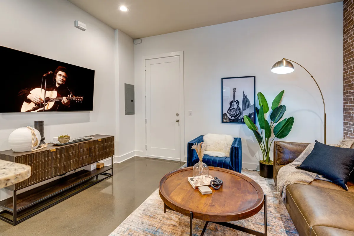 A cozy, stylish living room with a large flat-screen TV displaying a musician, a blue accent chair, a leather coffee table adorned with decor, and lush greenery from an indoor plant. The space is filled with natural light, highlighting the open-plan layout and contemporary design.