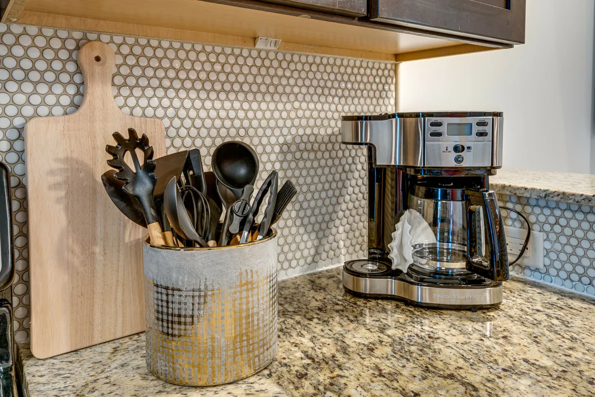 A kitchen close-up highlighting a coffee maker, utensil holder, and a cutting board on the countertop. The scene feels warm and approachable, suggesting easy meal prep and shared moments over morning coffee or snacks.