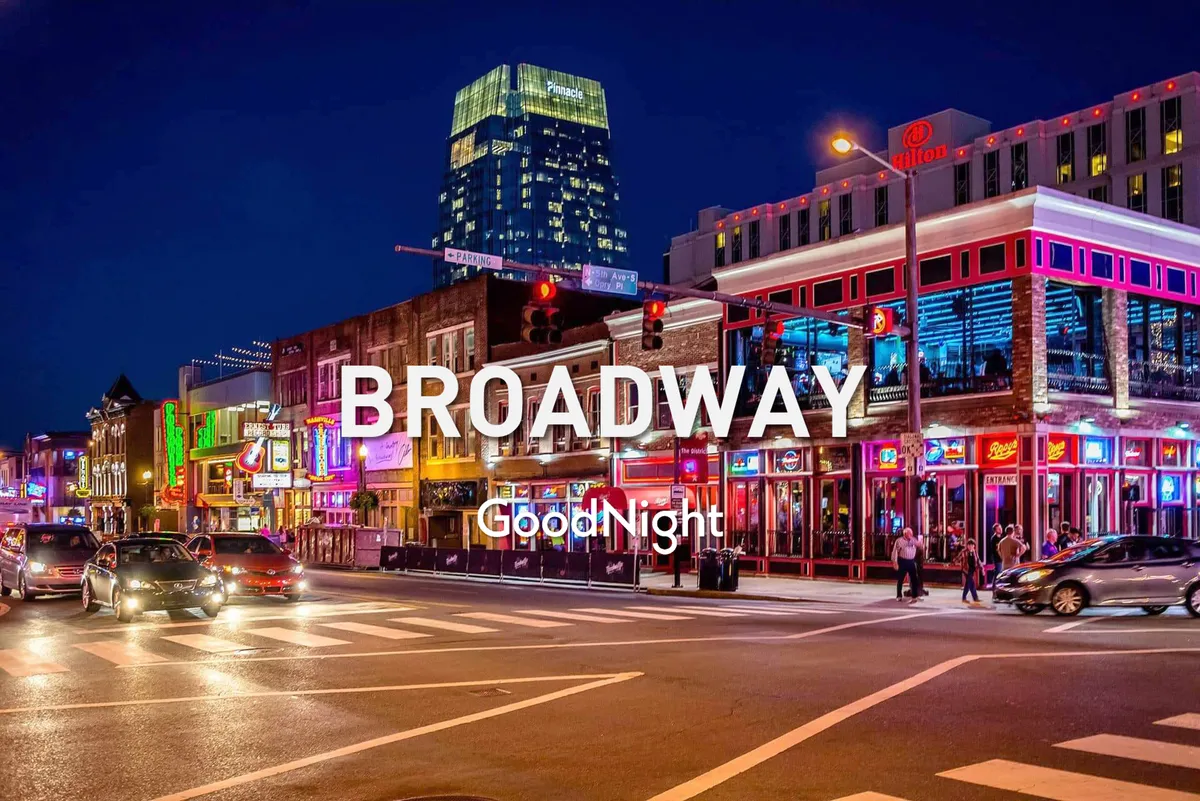 The image captures a lively urban streetscape at night, with neon signs glowing above a row of bustling restaurants and bars. People are seen walking and socializing, evoking the high-energy atmosphere of Nashville’s nightlife. The scene conveys a sense of excitement and vibrant culture, with traffic signals and crowds adding to the dynamic city feel.